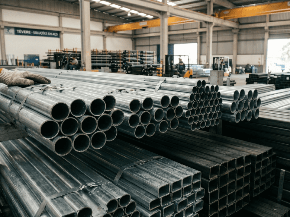 Close em estoque de tubos de aço galvanizado redondos e tubos de aço carbono quadrados organizados em prateleiras industriais dentro de um galpão logístico.