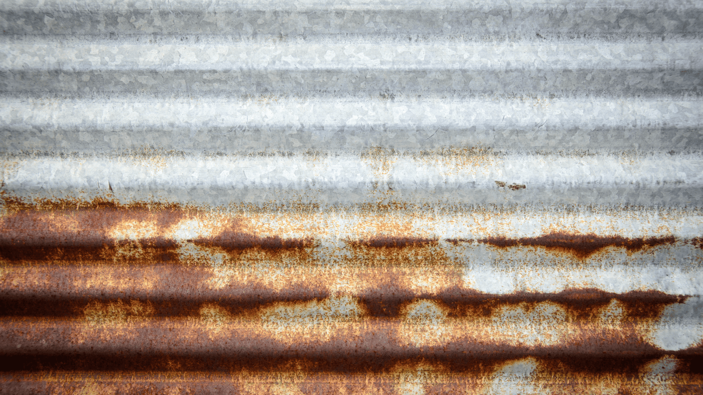 Textura de close-up de uma folha de metal ondulado horizontal, mostrando metal cinza-metálico limpo e galvanizado na metade superior e uma transição para ferrugem pesada, marrom e alaranjada na metade inferior.
