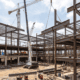 Canteiro de obras de um grande edifício comercial com estrutura de aço e concreto. Guindastes brancos e vermelhos operam sob céu nublado. Pilhas de materiais de metal no chão de terra.