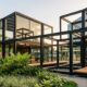 Fotografia de um pavilhão de arquitetura pop-up montado em um jardim. A estrutura modular é feita com perfis de aço preto aparente, panos de vidro e deck de madeira, sem pessoas no local. À direita, destaca-se um painel metálico preto com recortes vazados, exemplificando o uso de estruturas metálicas na construção efêmera